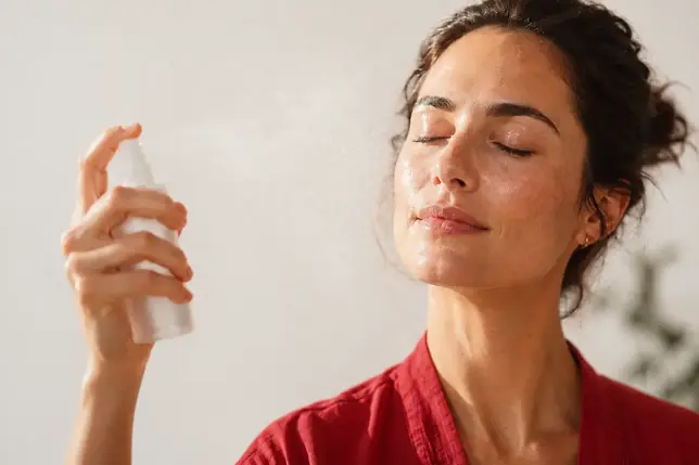 A woman misting a hypochlorous acid facial spray, eyes closed