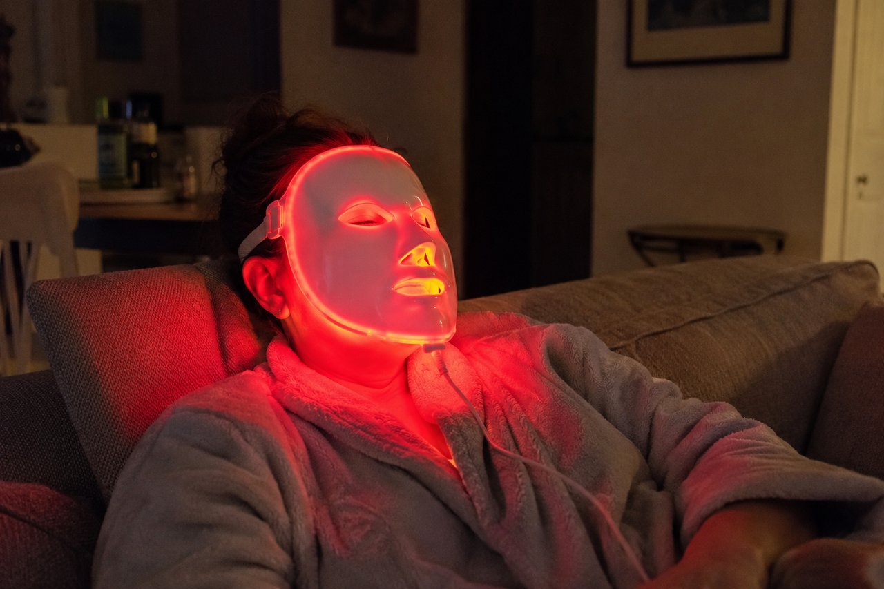 A woman relaxing on a couch wearing a glowing red LED light therapy face mask